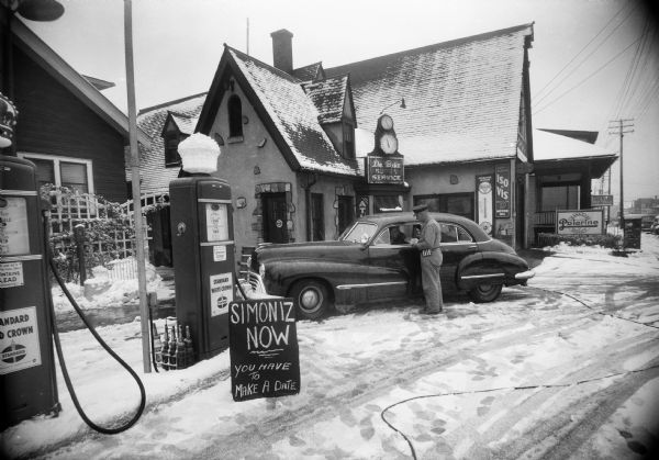A man stands next to an automobile in front of Du Bois Super Service at 1 North Mills Street (at the Corner of Regent Street), owned by Louis A. Du Bois. A sign advertises Simoniz service for cars.