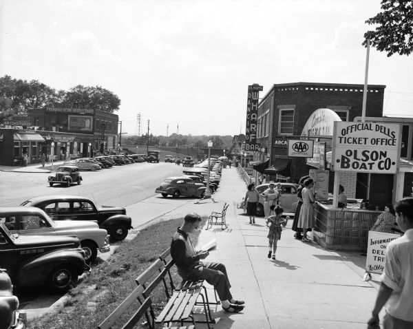 A view of downtown, including the official Dells ticket office. In the foreground a man is sitting on a bench reading a newspaper, and other pedestrians are walking along the sidewalk. There is a railroad bridge in the distance.