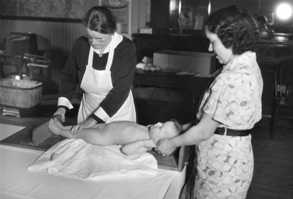 A nurse and a mother measure the length of a baby girl during an examination.