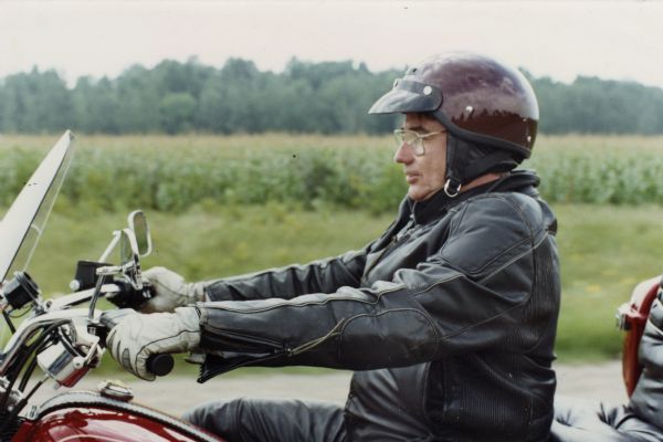 Wisconsin Governor Tommy G. Thompson wearing a leather jacket and riding a Harley-Davidson motorcycle on a rural road. A cornfield can be seen in the background.