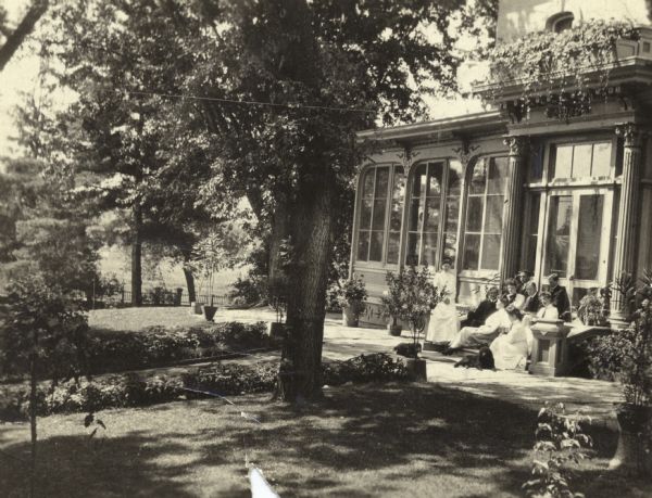 East facade view of Villa Louis with a group of four men, five women (possibly the Dousman family), and two dogs assembled on and around the steps at an entrance. A landscaped yard is visible in the foreground.