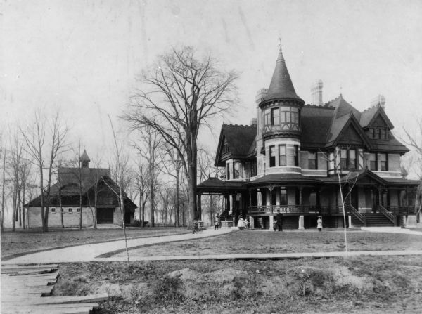 Exterior view of the Babcock home, a Queen Ann style structure at 537 E. Wisconsin Avenue. People, presumably the Babcock family, are standing in front of the house. A carriage house can be seen to the left of the home. A path leads from the house to the sidewalk in the foreground and a boardwalk is visible leading from the sidewalk to the street.
