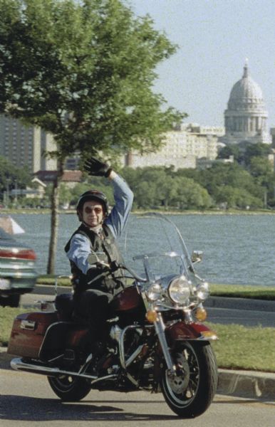 Wisconsin Governor Tommy G. Thompson waves from a Harley Davidson motorcycle as he rides on John Nolen Drive. Lake Monona and the Wisconsin State Capitol building are in the background.
