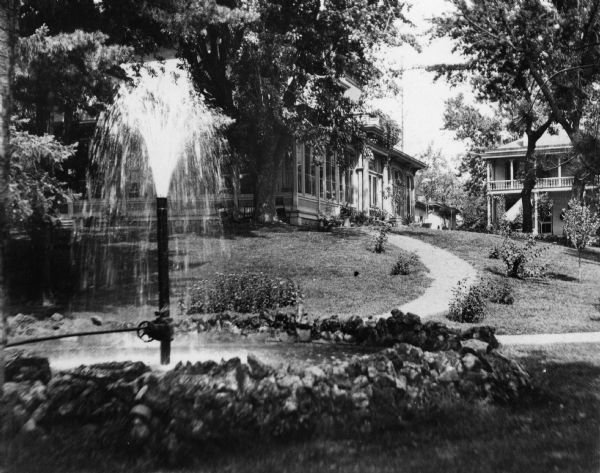 View of water spouting from a pipe sticking out of a pond surrounded by stones on the grounds of Villa Louis. The house and two outbuildings are visible among the trees in the background. A landscaped path leads from the house to the pond.