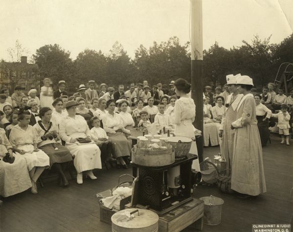 Group of women in U.S. Food Administration uniforms demonstrate food conservation methods to a group of men, women and children.
