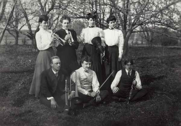 Group of four women standing and three men seated outdoors. They are all holding musical instruments except for the woman on the far right.