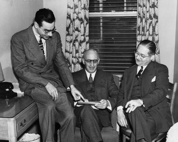 Elmer Berger, who is perched on the edge of a table, points to paperwork held by Lessing Rosenwald as I. Edward Tonkon looks on.