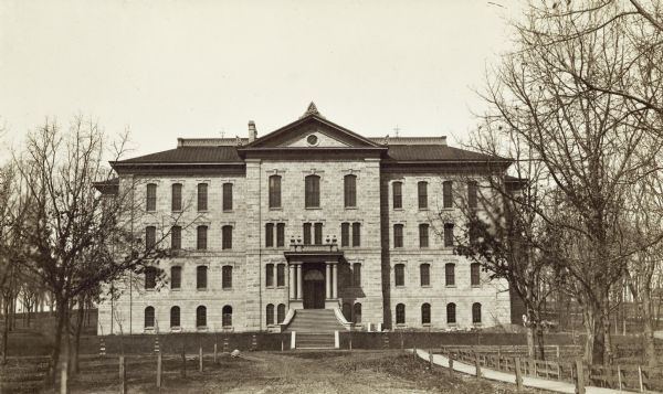 View down Langdon Street of building at base of Bascom Hill at the intersection with Park Street. This building stood from 1876-1884. A horse and carriage stand near the building on the right, behind a tree.