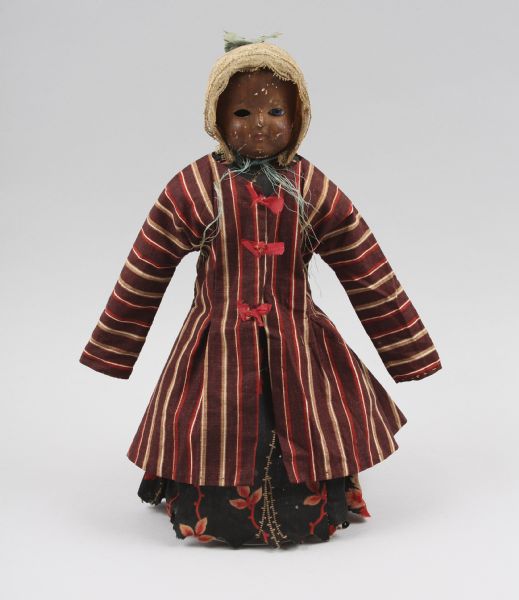 A girl doll dating back to 1890-1900. She is painted brown, and wearing a striped coat over a flowered dress.