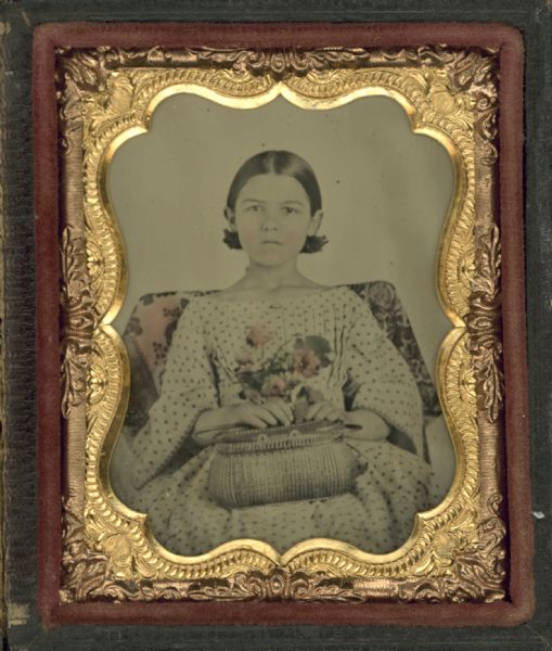 Ninth plate ambrotype of an unidentified young woman in dotted long sleeve dress, seated and holding a purse and a bouquet of flowers. Hand coloring on cheeks, flowers and chair. 