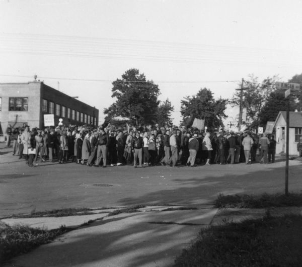 View from across street of a crowd of workers on strike, gathered in a parking lot near a railroad crossing.