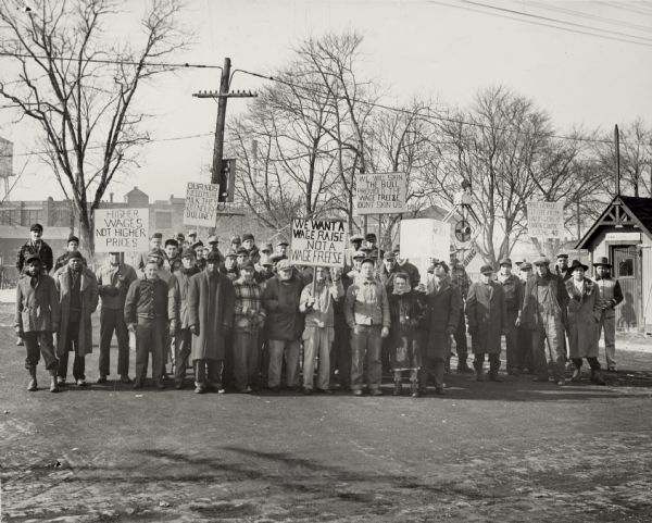 A group portrait near a railroad crossing of striking workers who hold signs with messages, including: "Higher Wages, Not Higher Prices".