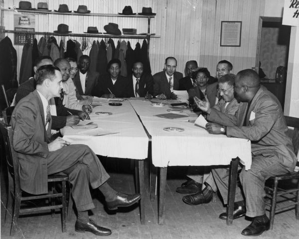 A formal meeting of men and women. All but one of those attending are African-American.