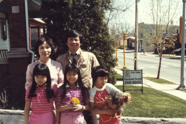 The Nguyen family pose for a family snapshot outdoors. A real estate sign that says "For Sale" is visible in the background in  a front yard. The parents, from left to right, are Hong and Trong Nguyen. The children, from left to right, are Mary, Thuy (holding dandelions) and Charlie Nguyen, who is holding a dog.