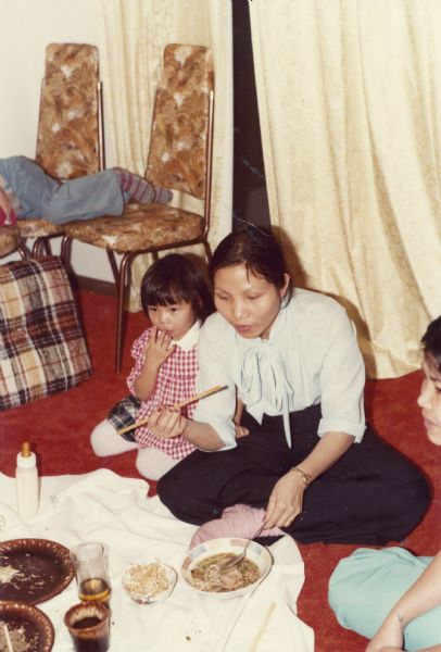 Hong Nguyen sits on the floor eating dinner. Her daughter Thuy, wearing a red and white dress, is sitting next to her, and an unidentified person is visible on the right.