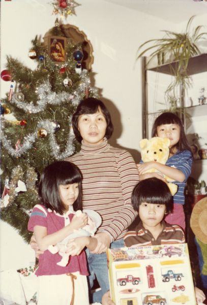 Hong Nguyen poses in front of a Christmas tree with three of her children. The children, from left to right, are: Mary, holding a doll, Minh, holding a box of toy trucks, and Thuy, holding a teddy bear.