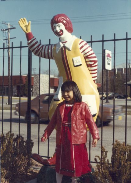 Thuy Nguyen poses in front of a Ronald McDonald figure in a public play area. She is wearing a red vinyl jacket. A Pizza Hut sign is visible in the background.
