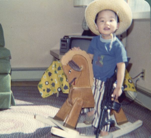 Minh Nguyen sits on a rocking horse. He is wearing a cowboy hat and holding a toy gun.