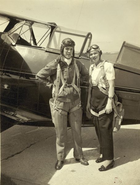 Dickey Chapelle, in a helmet and goggles, poses next to another pilot in front of an airplane.