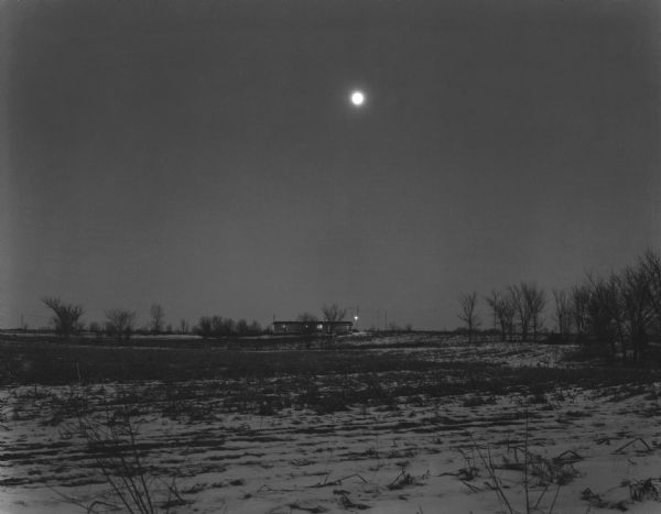 A ranch style farmhouse sits in the far distance beyond an empty field at night, with a full moon in a cloudless sky directly above.