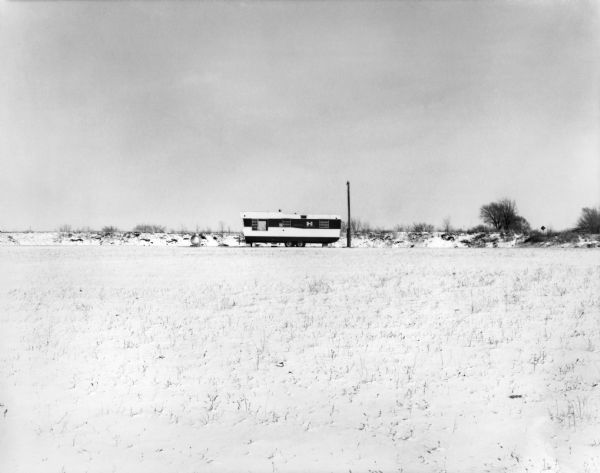 A mobile home sits alone at the edge of an empty field adjacent to a septic tank and telephone pole.