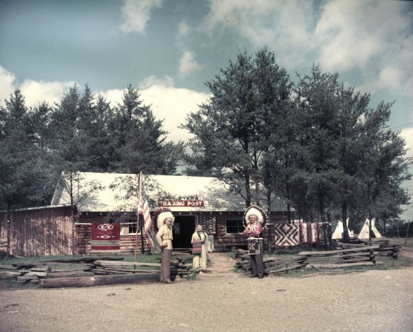 Two men and one woman in Native American costume stand near a fence and a U.S. flag outside the entrance of Pipe Dyer's Trading Post in Fort Dells. There are rugs hanging outside the store and teepees visible in the background. A sign on the trading post advertises film and postcards.