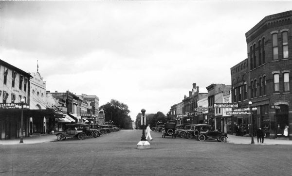 Downtown view. On the left side of the street is a restaurant, a photography store, a drugstore, and an ice cream parlor. On the right side is another ice cream parlor, the "Az-U-Lyk-It" Restaurant and Lunch Room, a clothes store, Lake Geneva Dry Goods, and a furniture store.