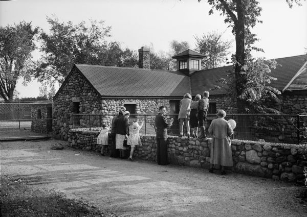 Several families peer into an animal environment at the Henry Vilas Zoo (Vilas Park Zoo). Three small children stand on top of the stone wall and look over the fence that encloses the area.