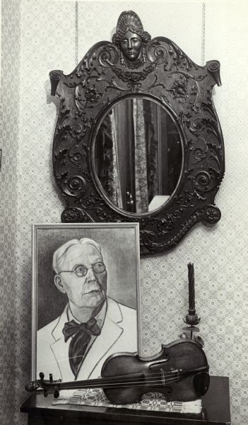Corner of room with a painting of Knute Reindahl, a violin, a candlestick and an ornate mirror, taken at Reindahl's home.