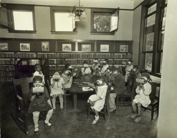 Group of children at the Cherry Street branch of the Evansville Public Library using steropticons to view stereograph images. Bookshelves line the back wall with art prints above them.