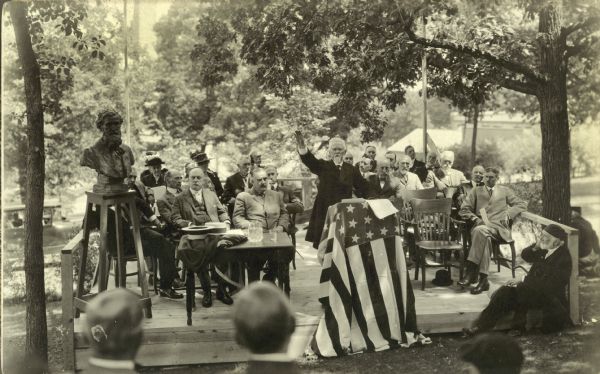 Dedication ceremony for Muir Knoll on the campus of the University of Wisconsin-Madison. Judge Milton S. Griswold is speaking at flag-draped podium, and a sculptured bust of John Muir, noted naturalist, stands on a pedestal on the left. Griswold was a friend of Muir.