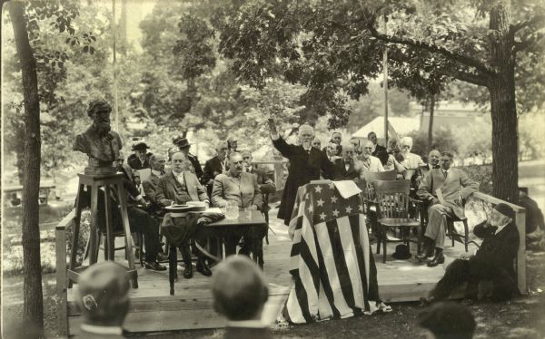 Dedication ceremony for Muir Knoll on the campus of the University of Wisconsin-Madison. Judge Milton S. Griswold is speaking at flag-draped podium, and a sculptured bust of John Muir, noted naturalist, stands on a pedestal on the left. Griswold was a friend of Muir.
