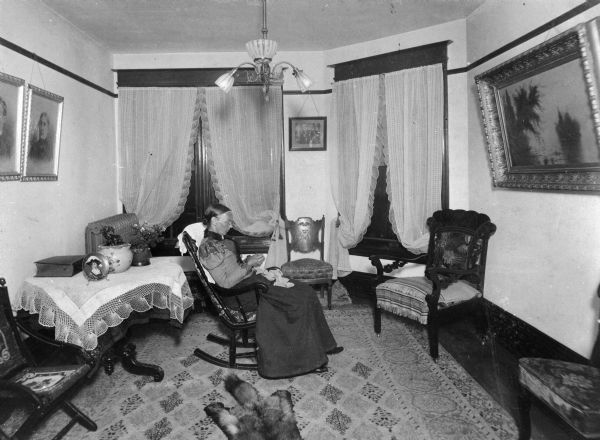 Mrs. Halle Steensland poses seated in a rocking chair in the beautifully decorated sitting room in her house at 315 North Carroll Street. She appears to be crocheting.