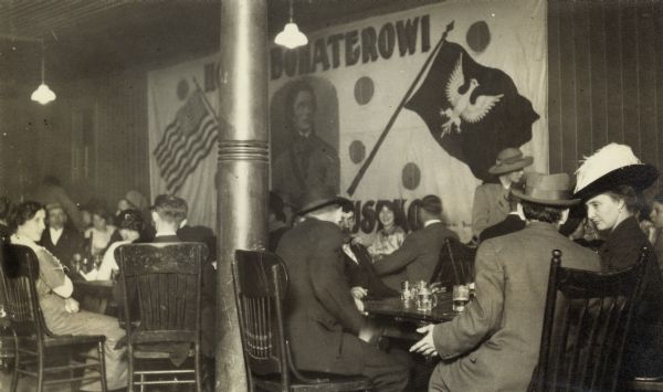 People sitting at tables, drinking and talking. A large banner with a picture of Thaddeus Kosciuszko hangs on the wall.