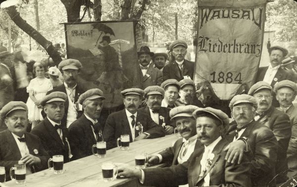 Members of the Wausau German Men's Choral Society in Sheboygan, gathered outdoors, relaxing and drinking beer. A banner behind the group says "Wausau Liederkranz 1884." The man who is seated third from the right is probably Jacob Kuhns.
