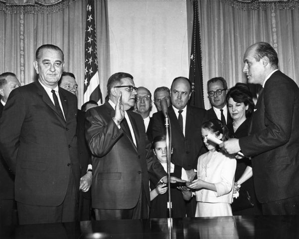 Attorney General Nicholas Kastenbach administers the oath of office to Postmaster General John A. Gronouski at the White House. From left to right are John Race (WI), President Lyndon Johnson, Whip Hale Boggs (LA), Gronouski, Lynn Stalbaum (WI), Senator Mike Maroney (OK), Melvin Laird (WI), Willard Wirtz, Mrs. Gronouski, and Katzenbach. In the foreground, Gronouski's daughters Julei and Stacy hold the Bible for the ceremony.