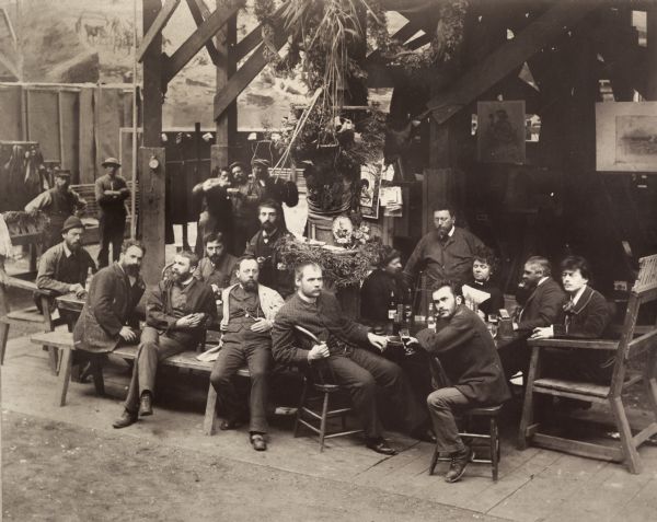 Group portrait of German painters relaxing in the studio of the American Panorama Company, during a break from painting the Jerusalem cyclorama depicting the crucifixion of Christ. Artists and their specialties include, at the left end of the table, Bernhard (Wilhelm?) Schroeder (Schroeter) wearing a hat (landscapes), and behind the table from the left, Richard Lorenz (animals), Johannes Schulz (figures), Friedrich Wilhelm Heine (Supervisor and master of composition), Bernhard Schneider (landscapes), Irma (Amy?) Boos, and August Lohr in profile (Supervisor and designer of landscape settings). Seated in front of the table are Franz Bilberstein (landscapes), Franz Rohrbeck (figures, especially Confederate), Karl (Carl) Frosch (Frosh), Thaddeus Zukotynski (Zuchatinsky, Chuchodinski) (figures), George Peter (animals), and Herman Michalowski (figures).