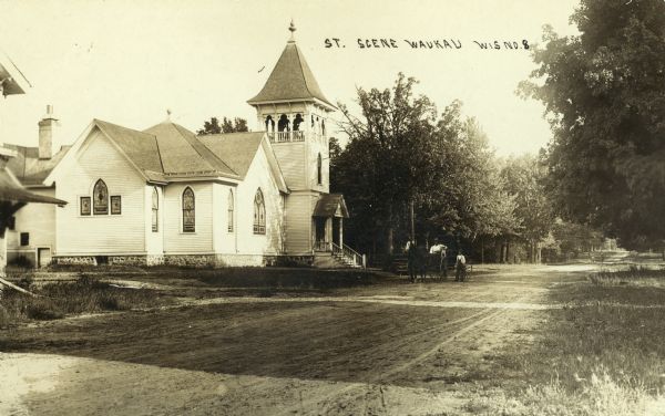 Street scene featuring a Methodist church. In front of the church a young boy stands in the dirt road near a man sitting in a horse-drawn wagon with a dog. Caption reads: "St. Scene Waukau Wis."