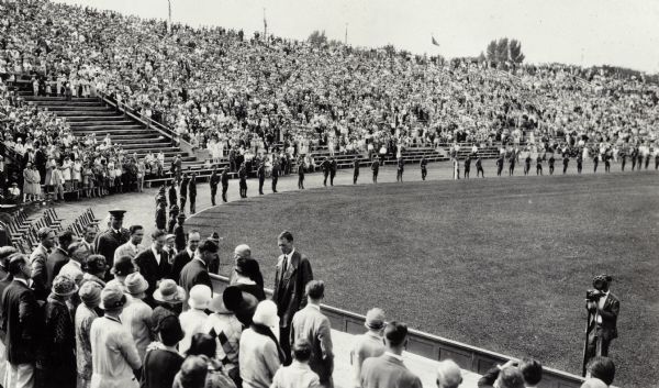 Charles Lindbergh meeting local dignitaries, with the crowd of admirers behind him seated in the stadium. Partly obscured to his right is Governor Schmedeman and further to the left, Secretary of State Fred Zimmermann.