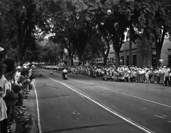 Madison Soap Box Derby view of the crowd watching the race in front of Water Works, corner of East Gorham and North Hancock Streets. Motorcycle Officer, C.J. Sparks, follows two racers down the hill on East Gorham Street.
