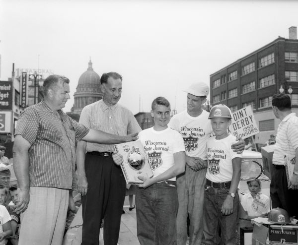 Van Steiner, winner of the 1957 Madison Soap Box Derby, holding the trophy. Left to right: Larry Steiner, father of the winner; Don Knowles, general manager of Hult Capitol Garage, a co-sponsor of the race; Wally Wikoff of the Wisconsin State Journal, also a race co-sponsor; and Jim Van Epps, the class B winner. Steiner of Argyle went on to win the national competition. Visible in the background (at left) is the Firestone Tire Co., 215 East Washington Avenue, and (at right) is the Ray-O-Vac Office, 212 East Washington Avenue.  The Wisconsin State Capitol stands in the middle.
