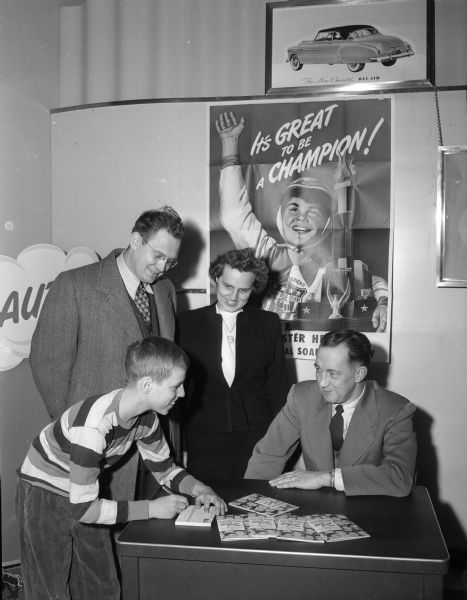 Chuck Dykman, shown with his parents Melvin and Ruth Dykman, signs up for the 1950 Soap Box Derby at Hult's Chevrolet garage.  Don Knowles, vice-president at Hult's is seated at the table at right. A poster for the Soap Box Derby hangs on the wall behind the group.