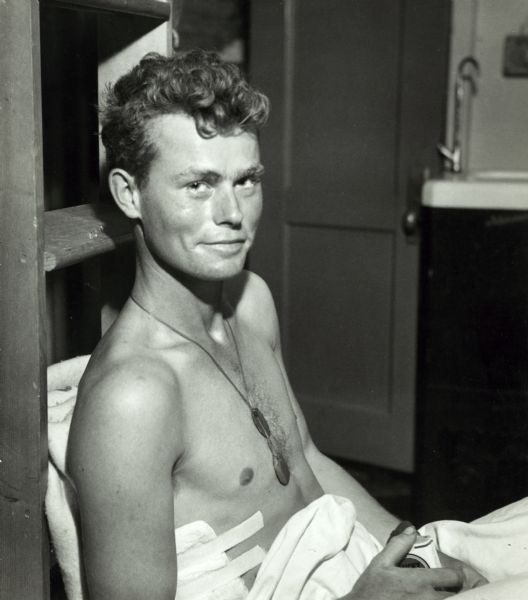 Private first class, John W. Hood, wearing dogtags smiles after a blood transfusion on the USS <i>Samaritan</i> in Iwo Jima. He is bandaged and is holding a pack of Lucky Strike cigarettes.