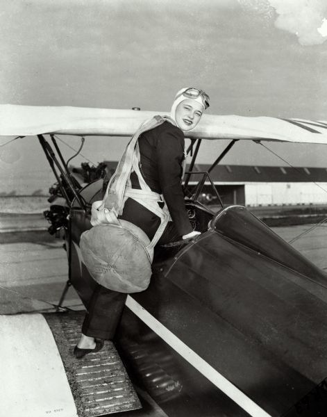 Dickey Chapelle getting into a small airplane. She is wearing a pilot's helmet, goggles and a scarf, and is carrying a parachute.