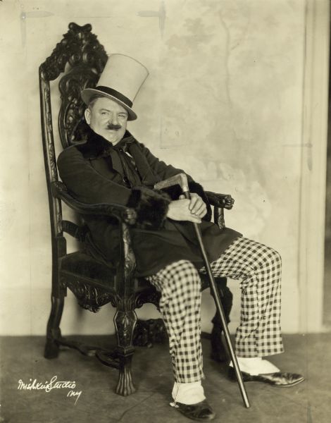 W.C. Fields in a publicity still from the Broadway show "Poppy". He is seated in an ornate chair and is wearing a top hat, checkered pants and spats.
