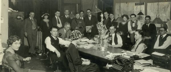 Journalist Robert S. Allen, center, in military uniform, poses with the staff of the "Wisconsin State Journal". A graduate of the Wisconsin Journalism School, Allen worked for both the "Capital Times" and the "Wisconsin State Journal" before moving on to national prominence as a syndicated columnist. There is a small Christmas tree on the table that people are gathered around.