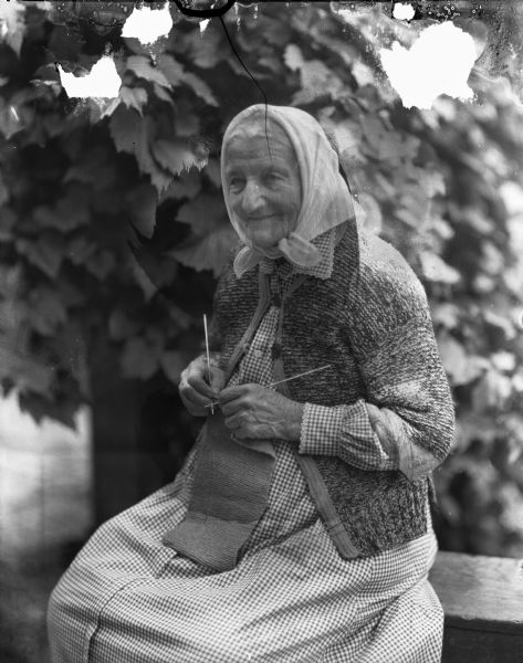 An elderly woman in headscarf, checked dress and sweater, sits outdoors knitting. Identified as Martha Schilling, wife of Adam Rebholz from Portage, Wisconsin.