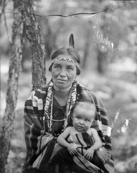 A Native American woman wearing a mix of traditional and western dress holds a child in her lap outdoors.