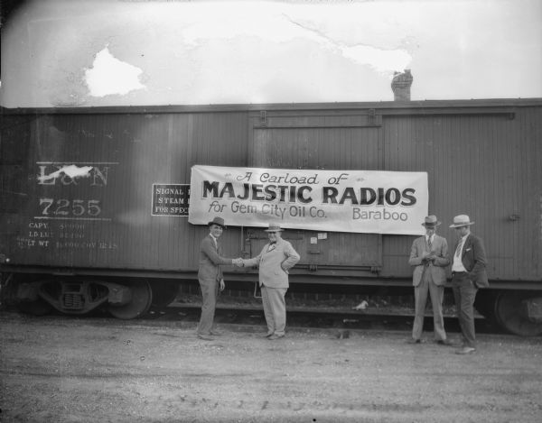 Four men pose in front of a railroad car. Two of the men are shaking hands. A banner on the car advertises "A Carload of Majestic Radios for Gem City Oil Co. Baraboo." The railroad car is marked "L & N," for the Louisville and Nashville Railroad.