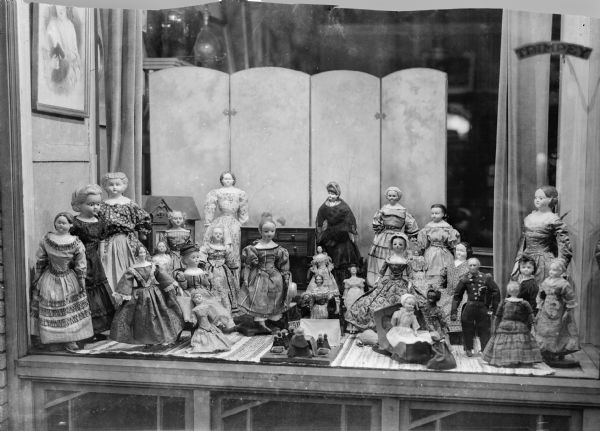 Dolls from the collection of Alice Kent Trimpey arranged in the front window of the Trimpey Studio and antique shop at 128 Fourth Avenue.  There is a miniature dresser and dollhouse in the background.